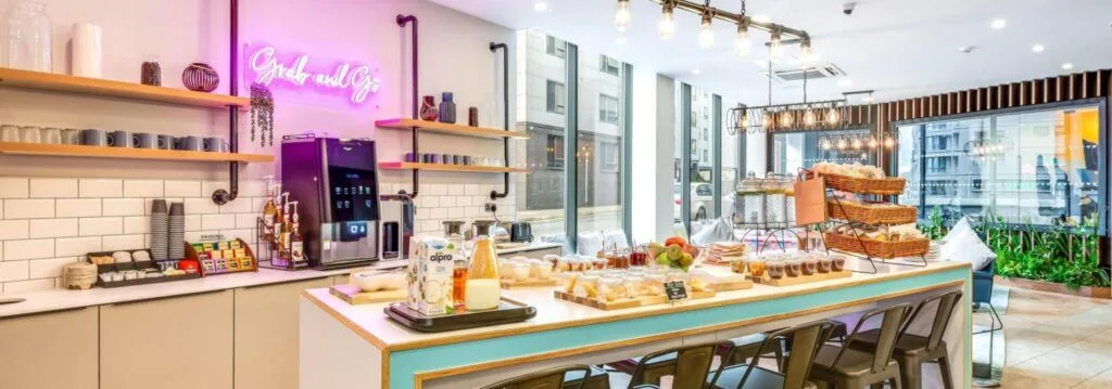 A bright and colourful breakfast bar and table full of food and drinks including pastries, fruit, milk, coffee and tea, inside a student accommodation building in Leeds. 