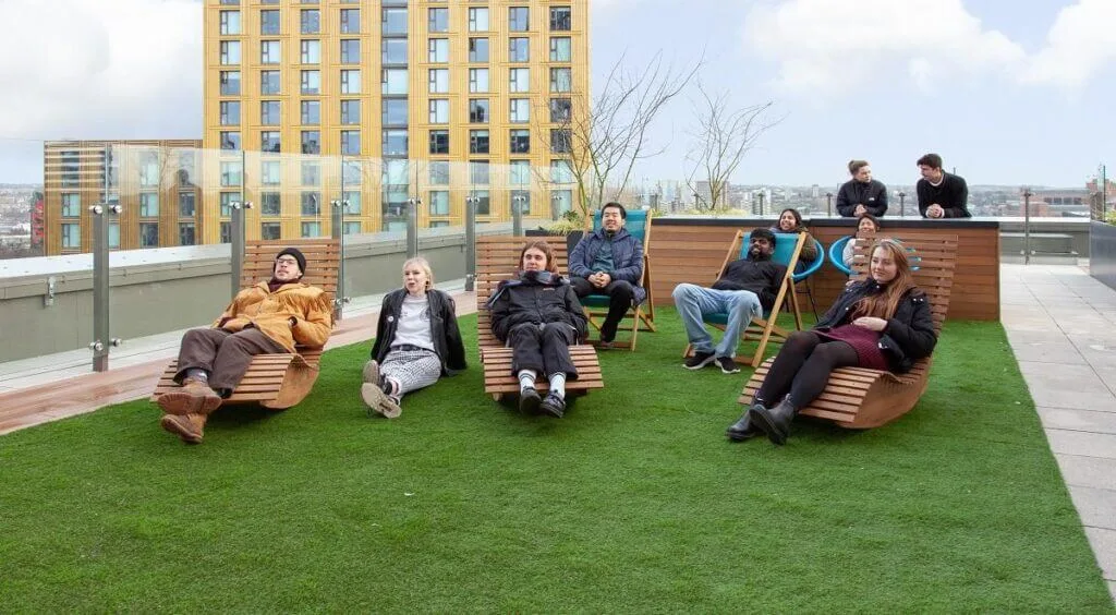 A student accommodation building's rooftop with wooden chairs, Astro turf, and ten students sitting in their winter clothes on a winter's day in Leeds. 