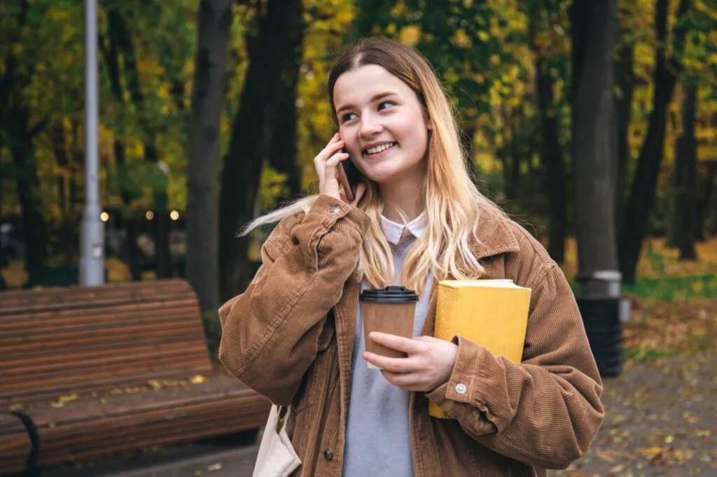 A student holding a coffee and a book while chatting on the phone in a park in autumn