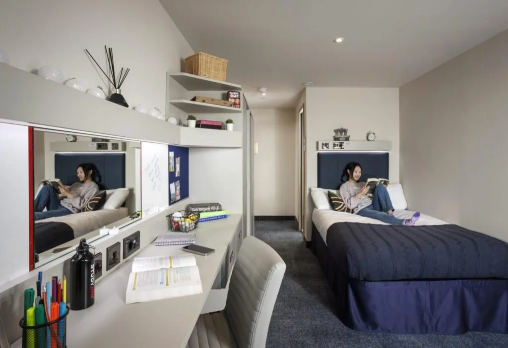 A student reading on her bed in an en-suite room at Study Inn Brotherton House, Leeds, with a desk, shelves and a pin board on the wall