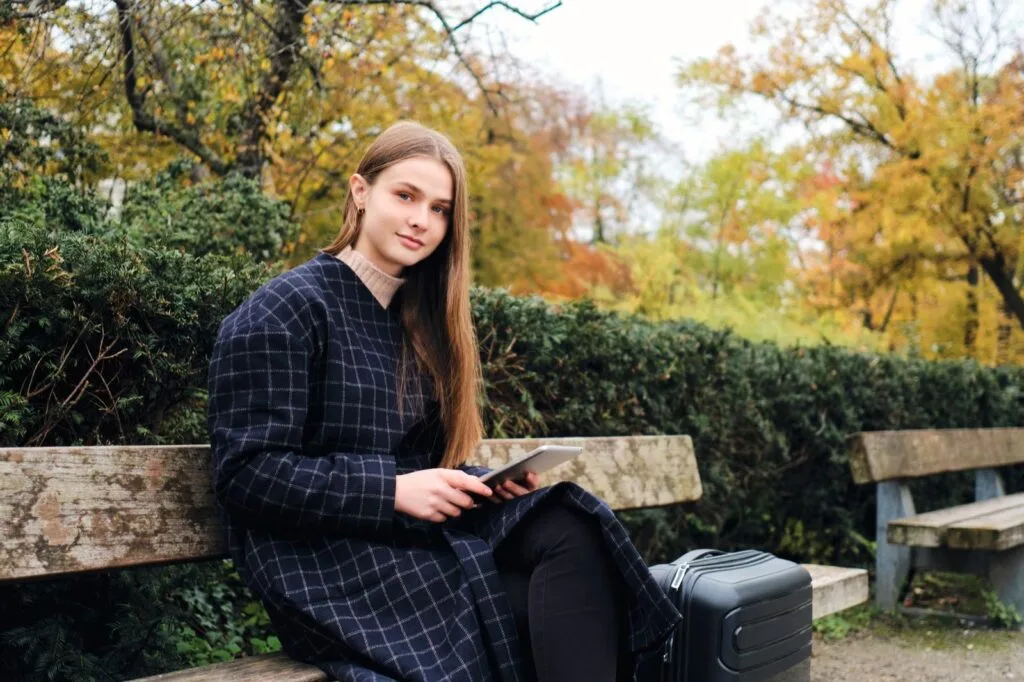 A student with a suitcase sitting on a park bench and checking a tablet